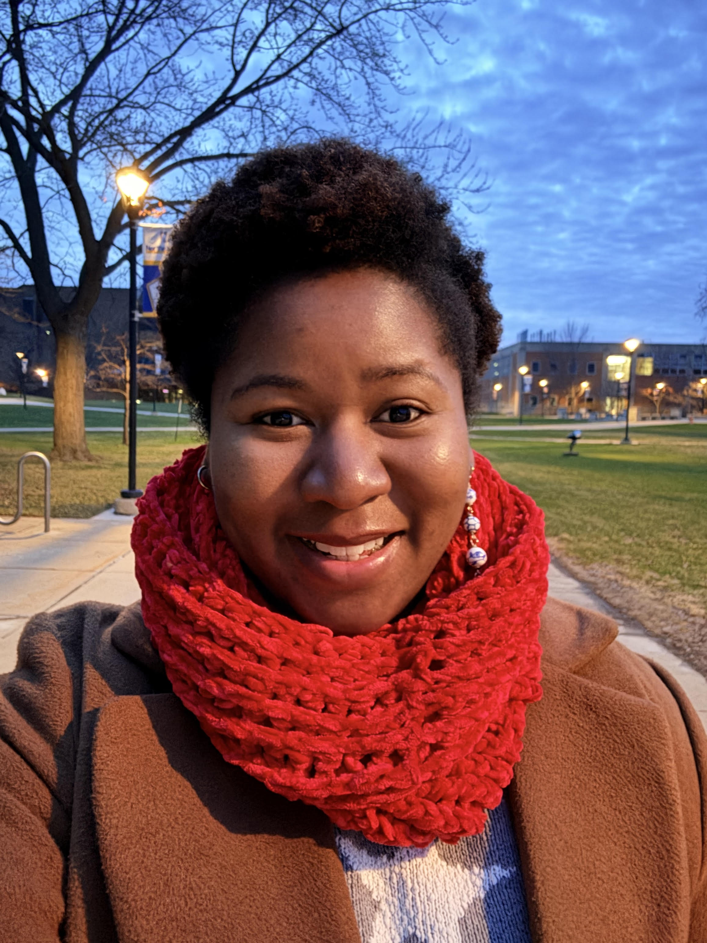 Yemilee Lempa, a smiling woman with dark hair wearing a tan jacket and red textured scarf, photographed at dusk on a university campus quad. Behind her are bare trees illuminated by warm street lamps and campus buildings glowing with interior lights. Her warm, genuine smile and calm confident expression convey approachability and professionalism.